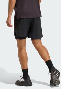 Pantalones cortos deportivos negros con un diseño ligero, que presentan aberturas laterales, superpuestos sobre pantalones cortos negros ajustados. Calcetines y zapatos negros completan el conjunto.