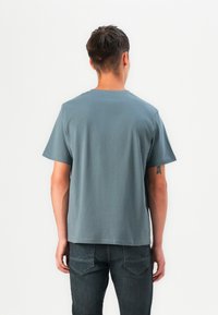 Tricou din bumbac gri, cu guler rotund și mâneci scurte, purtat cu blugi negri. Design simplu, fără modele sau grafice vizibile.