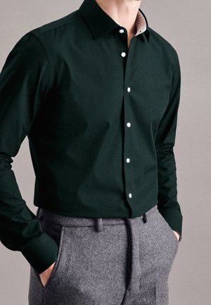 Man in een donkergroen, knoopshirt, gestoken in grijze wollen broeken, met handen in zijn zakken, staande tegen een eenvoudige neutrale achtergrond.