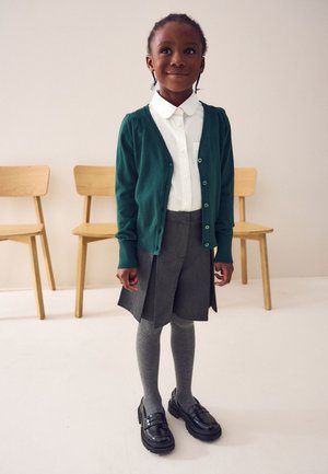 Mladé dievča v školskom uniforme s zeleným kardigánom, bielou košeľou, sivou sukňou a pančuchami stojí pred drevenými stoličkami na bielom podlaží.