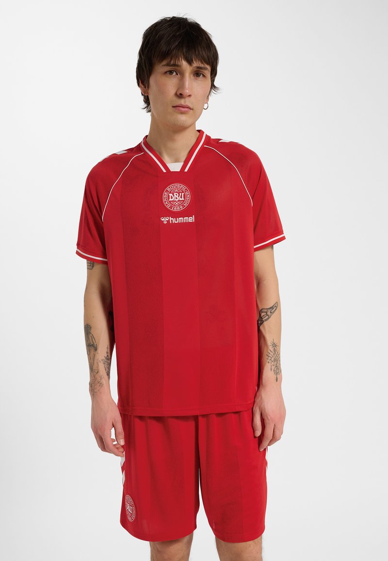 Giovane uomo con capelli corti e scuri che indossa una maglia rossa della nazionale di calcio danese e pantaloncini abbinati, in piedi davanti a uno sfondo semplice.