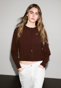 Burgundský zkrácený cardigan s kulatým výstřihem, žebrovanou texturou a černými knoflíky, v kombinaci s bílými kalhotami s vysokým pasem.