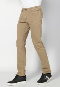 Pantalones de algodón beige con corte ajustado, diseño de cinco bolsillos y costuras en contraste. Llevados con zapatillas negras sobre un fondo blanco.