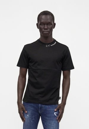 Homme portant un T-shirt noir à manches courtes avec un texte blanc près du col et un jean bleu déchiré, debout devant un fond blanc.