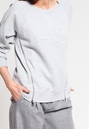 Persoon draagt een lichtgrijze trui met twee verticale ritsen aan de voorkant aan de zijkanten en bijpassende grijze broek, handen in de zakken.