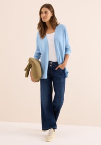Vrouw in een lichtblauwe cardigan, witte top, wijde donkere jeans en beige sneakers, met een opgevouwen bruine jas in één hand.