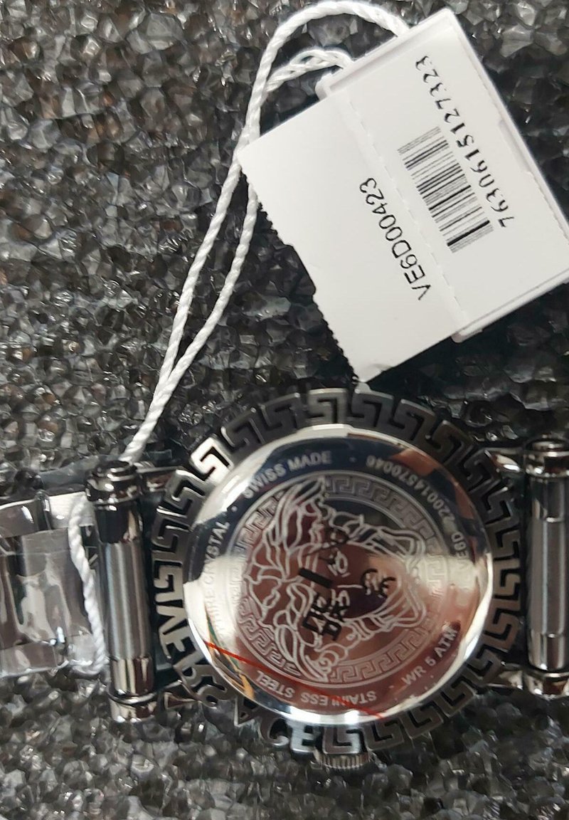 Montre en acier inoxydable argenté avec un design de clé grecque, présentant une gravure de lion au dos et une étiquette de prix accrochée.