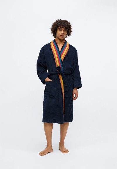 Mees, kes seisab paljajalu, seljas tumesinine hommikumantel mitmevärvilise triibulise äärise ja vööga, käed taskutes, neutraalse taustaga.