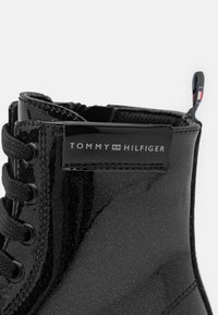 Zwarte lakleren enkelboot met zwarte veters, voorzien van een zilveren glans en een Tommy Hilfiger logo tag aan de zijkant.
