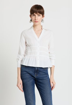 Chemise blanche à boutons avec une taille smockée, des manches longues et un design péplum, associée à un jean en denim bleu.
