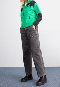 Vihreä ja musta nailonjacket, jossa on vetoketjulliset taskut, yhdistettynä tummanharmaisiin cargo-housuihin, joissa on sivutaskut. Mustat nilkkasaappaat, joissa on teksturoitu pohja.