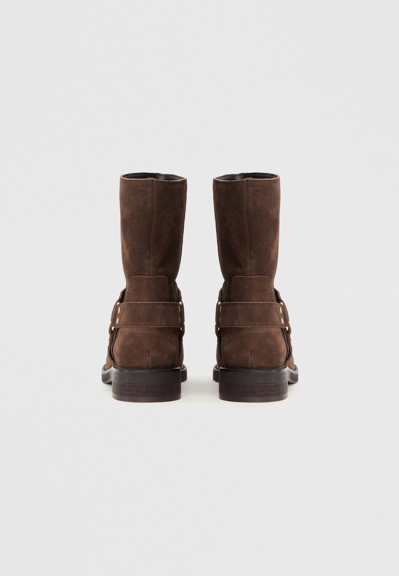 Braune Wildlederstiefel bis zur Mitte der Wade mit einer glatten Oberfläche, die über eine Schnalle und eine robuste schwarze Sohle verfügen. Einfache, klare Linien betonen das Design.