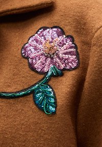 Καφέ ένδυμα από τσόχα με ροζ λουλουδάτο ύφασμα με παγιέτες, που περιλαμβάνει πράσινα φ ύλλα με παγιέτες και μαύρο περίγραμμα, δημιουργώντας μια υφασμάτινη σχεδίαση.