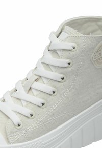 Baskets montantes en toile beige clair avec des lacets blancs, une tige cousue et une semelle en caoutchouc texturée avec un accent à motifs.