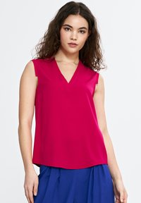 Fuksjowa bezrękawnikowa bluzka z dekoltem w kształcie V, wykonana z gładkiego materiału, z zaokrąglonym dołem i luźnym krojem, zestawiona z niebieskimi spodniami.