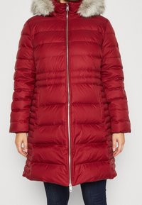 Vrouw in een lange, rode gewatteerde winterjas met een zilveren rits aan de voorkant en een beige capuchon met bontafwerking, met haar handen langs haar zij.