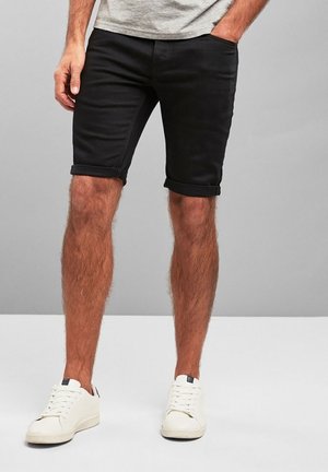 Czarne dżinsowe szorty z podwiniętymi nogawkami, kieszeniami z przodu i dopasowanym krojem, w połączeniu z białymi trampkami na szarym tle.