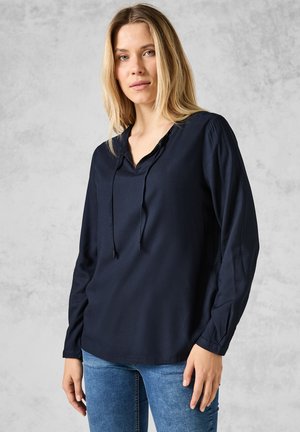 Camisa de manga larga azul marino, fabricada en un tejido ligero, con escote en V y cordón ajustable, y mangas fruncidas. Se lleva con jeans azules sobre un fondo neutro.