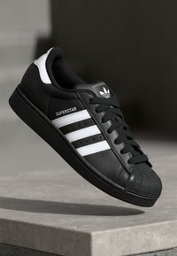 Black leather sneaker with three white stripes, rubber shell toe, black laces, and a logo patch on the tongue. Lightweight design.