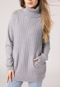 Jasnoszary sweter w splot kablowy z wysokim golfem, ściągaczami przy rękawach i kieszeniami z przodu. Miękka tekstura i luźny krój.