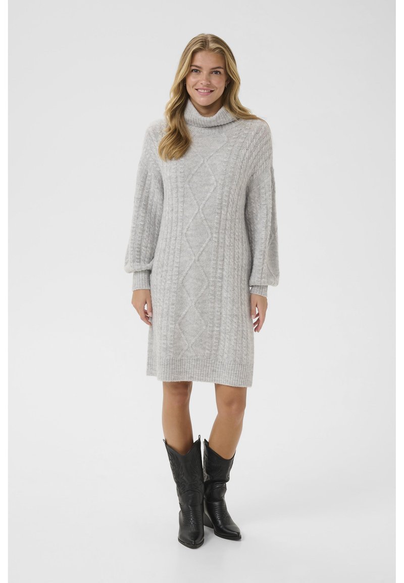 Grå stickad klänning med hög krage, prydd med texturerade flätmönster och långa ärmar, kombinerad med svarta cowboy boots.