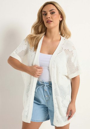 Kobieta w białym, luźnym kardiganie z krótkim rękawem na białym topie na ramiączkach i jasnoniebieskich szortach ze ściągaczem, stojąca na jednolitym tle.