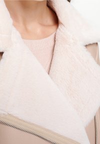 Giacca di pelliccia sintetica rosa chiaro con colletto ampio, dotata di una zip beige liscia e una consistenza morbida, indossata sopra un maglione rosa pallido.