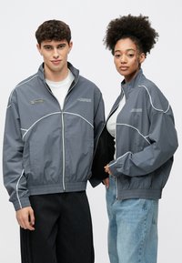 Grå hettejakker med glidelås og hvite detaljer, med høy krage og elastiske mansjetter, båret av to modeller. Stoffet virker lett og glatt.