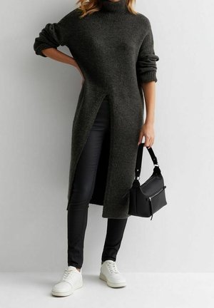 Femme portant un long pull gris foncé avec une fente à l'avant, un pantalon noir ajusté, des baskets blanches et tenant un sac à main noir.