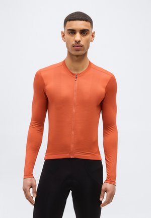 Νέος άνδρας που στέκεται αντικρίζοντας μπροστά, φορώντας στενή ποδηλατική μπλούζα μακριάς μανίκας σε πορτοκαλί χρώμα και μαύρο ποδηλατικό σορτς, μπροστά σε απλό φόντο.