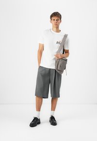 Hvit bomulls t-skjorte med "REPLY" trykk, grå plisserte shorts, svarte sko, grå skulderveske, står mot en ensfarget bakgrunn.
