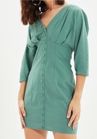 El vestido verde de manga corta presenta un escote en V, un cuerpo plisado y un cierre frontal con botones a juego, fabricado con una tela suave.