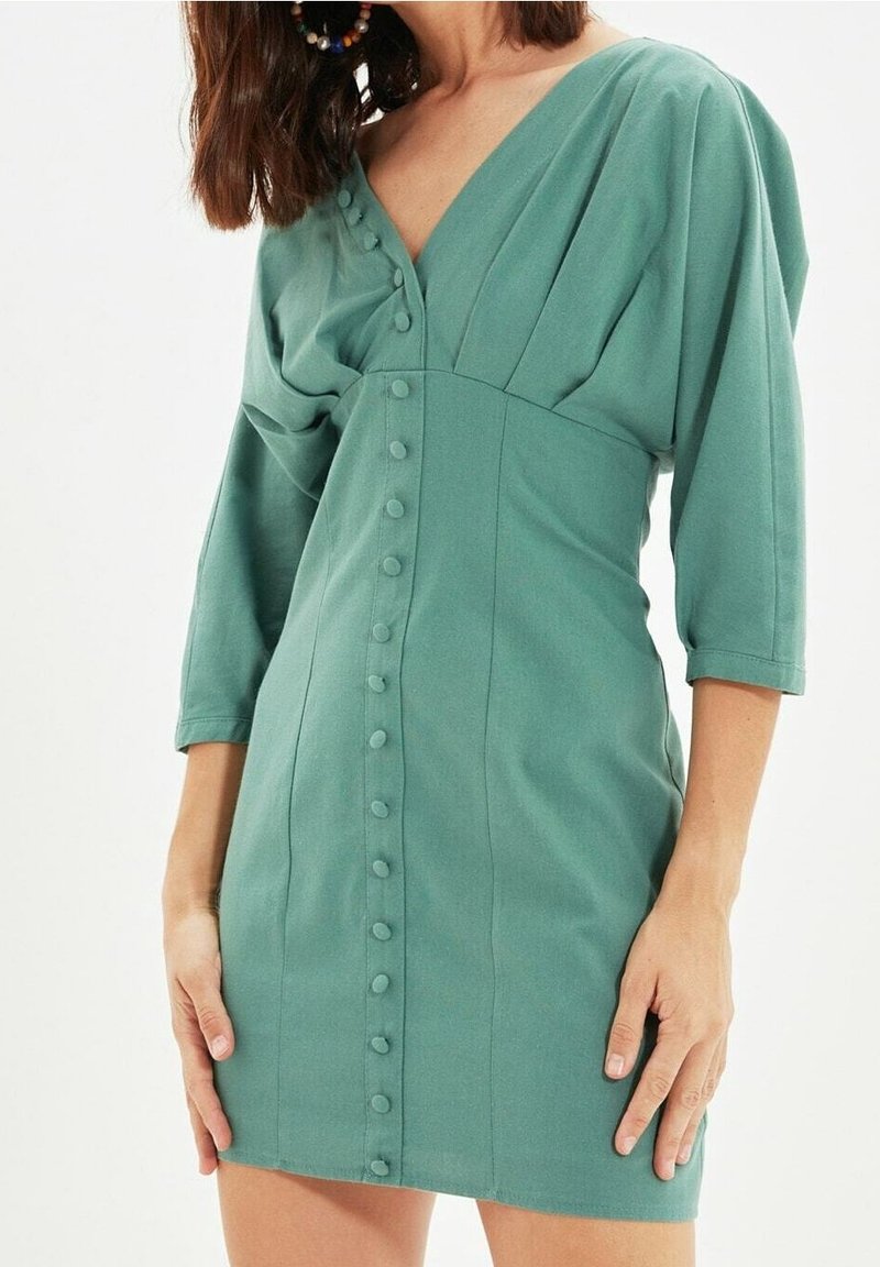 El vestido verde de manga corta presenta un escote en V, un cuerpo plisado y un cierre frontal con botones a juego, fabricado con una tela suave.