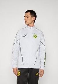 Veste de sport zippée blanche avec des accents noirs et un logo vert. Présente un col haut et des designs de panneaux latéraux. Assortie avec un pantalon noir.