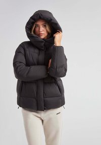 Czarna kurtka puchowa z kapturem, o pikowanej konstrukcji, z zapięciem na zamek z przodu i elastycznymi mankietami. Nosi się ją na jasnobeżowe spodnie dresowe.