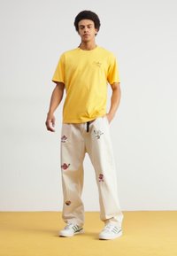 Geltonas medvilninis marškinėliai ir šviesiai kreminės kelnės su išsiuvinėtais spalvingais raštais. Baltos sportinės bateliai su žaliaisiais dryželiais. Modelis stovi prieš neutralų foną.