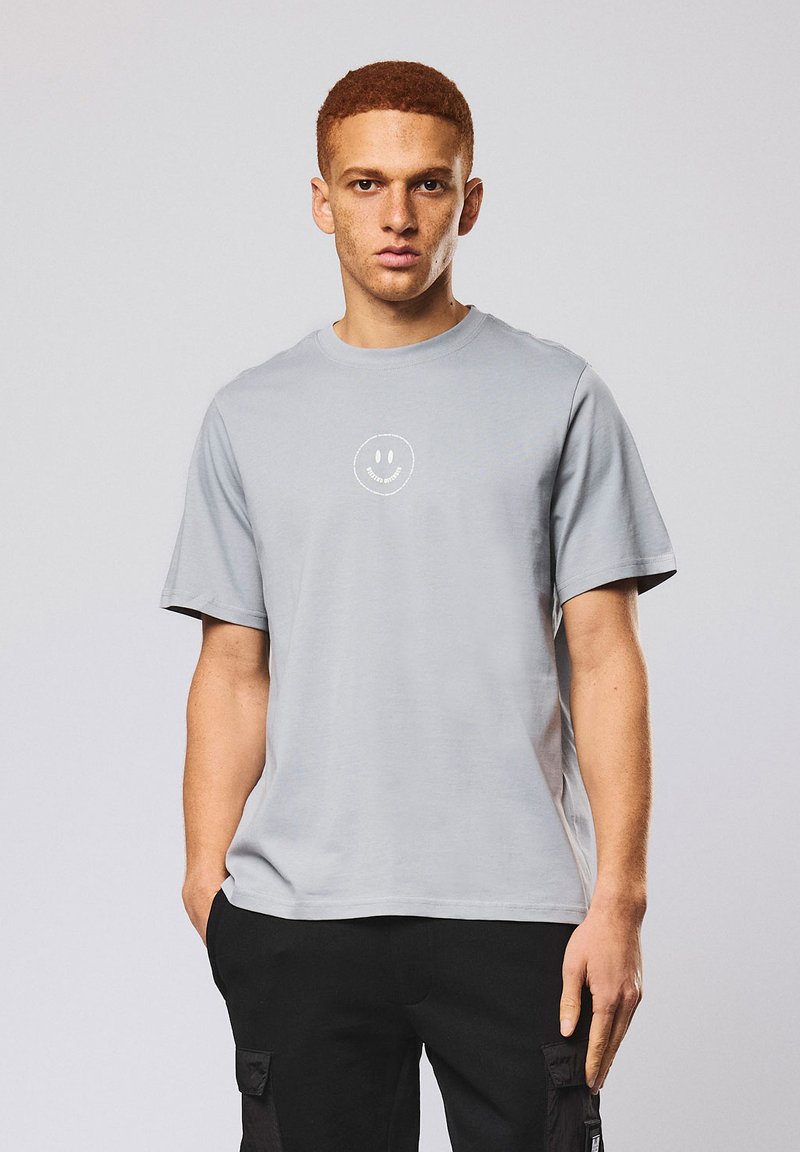 Jeune homme aux cheveux roux courts portant un T-shirt gris clair avec un petit smiley blanc et un pantalon cargo noir, debout les mains dans les poches.