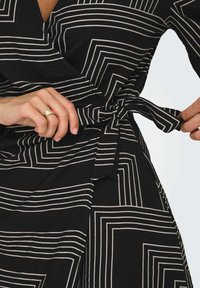 Crna haljina na preklop sa bijelim geometrijskim linijama, teksturiranim materijalom i pojasom koji se veže. Kratki snimak ruke koja drži kravatu.
