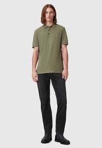 Mladý muž stojí oblečený v olivovo zelenej polokošeli s krátkym rukávom, čiernych džínsoch a čiernych kožených čižmách na jednoduchom pozadí.