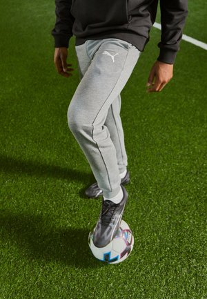 Person trägt graue Puma-Sweatpants und schwarze Fußballschuhe und kontrolliert einen Fußball auf grünem Kunstrasen.