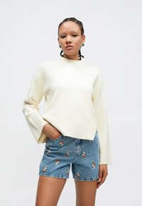 Kremfarget, oversized genser med høy hals og ribbestrikket kant, kombinert med lyseblå denimshorts med blomsterbroderi.