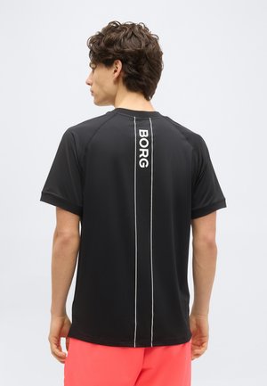 Νεαρός άνδρας που φοράει μαύρο μπλουζάκι με λευκές κάθετες ρίγες και το κείμενο "BORG" στην πλάτη, συνδυασμένο με φωτεινά κοραλλί σορτς.