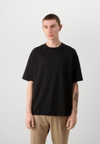 Black short-sleeve t-shirt made of cotton featuring an embossed "Calvin Klein" logo on the front. Relaxed fit, round neckline.