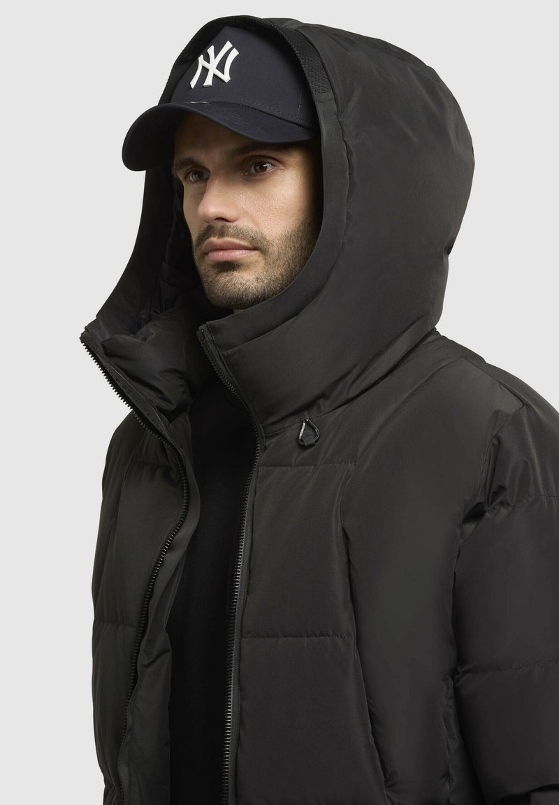 Schwarze Pufferjacke mit großer Kapuze und Reißverschluss, die eine glatte Textur und einen Knopfakzent aufweist. Getragen mit einem kontrastierenden schwarzen Oberteil und einer Mütze.