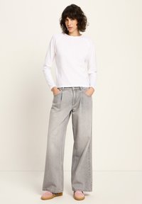 Frau steht mit den Händen in den Taschen, trägt ein weißes Langarmoberteil, hellgraue weite Jeans und rosafarbene Turnschuhe vor einem einfarbigen Hintergrund.