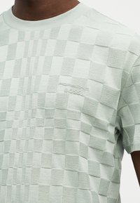 Ανοιχτό πράσινο κοντομάνικο πουκάμισο με υφή καρό μοτίβο και ανάγλυφο λογότυπο "Porsche BOSS" στο στήθος, φορεμένο από ένα άτομο.