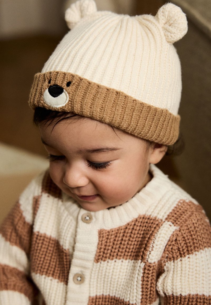 Bonnet tricoté à thème ours avec une texture côtelée crème et beige, comportant des détails du visage d'ours. Porté avec un cardigan assorti à rayures.