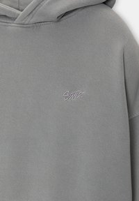 Sweat à capuche gris clair en tissu doux, avec un petit logo brodé blanc sur la poitrine. Design simple avec une coupe décontractée.
