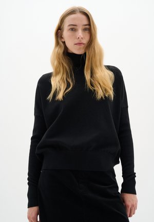 Schwarzer Rollkragenpullover aus weichem Stoff, mit lockerer Passform und langen Ärmeln. Die Silhouette wird mit dunklen Hosen kombiniert.