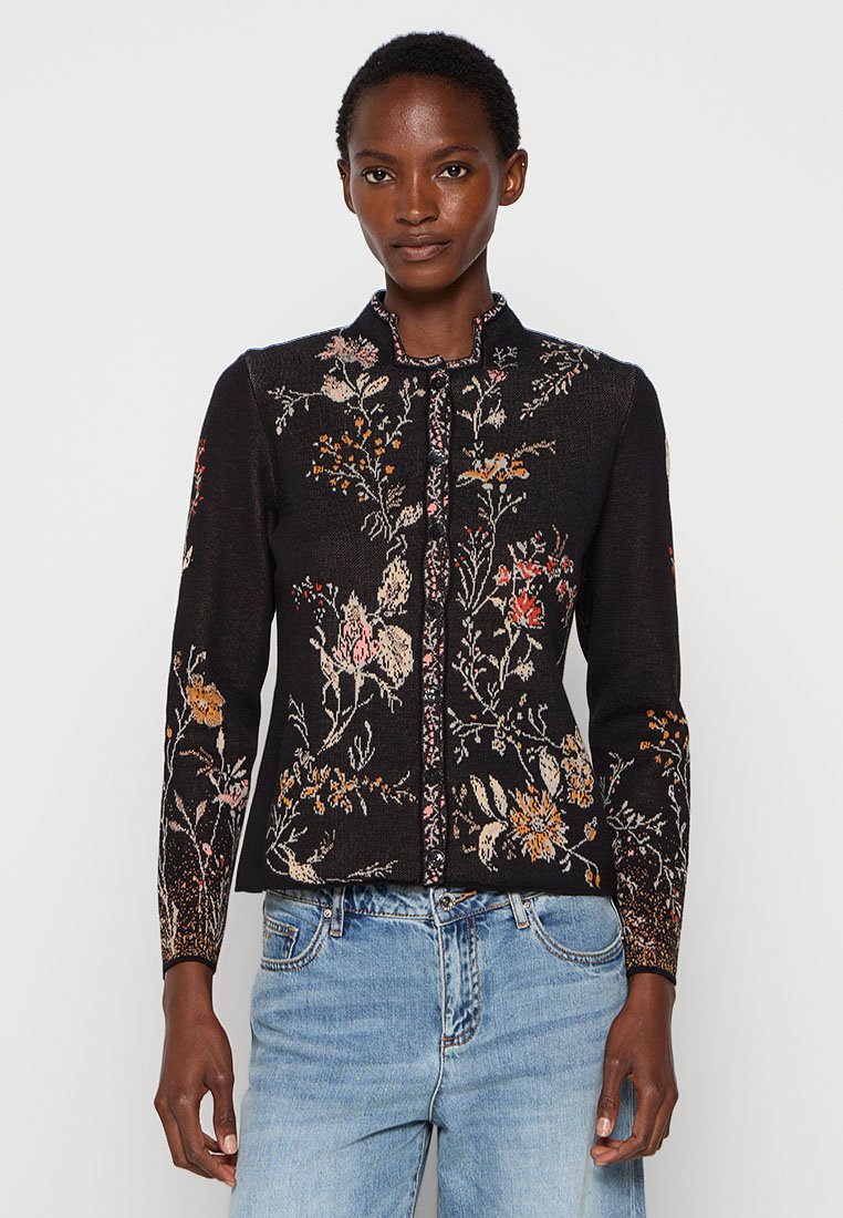 Vrouw in een zwarte cardigan met bloemenpatroon en lange mouwen, met een staande kraag en lichtblauwe jeans, staande tegen een effen achtergrond.